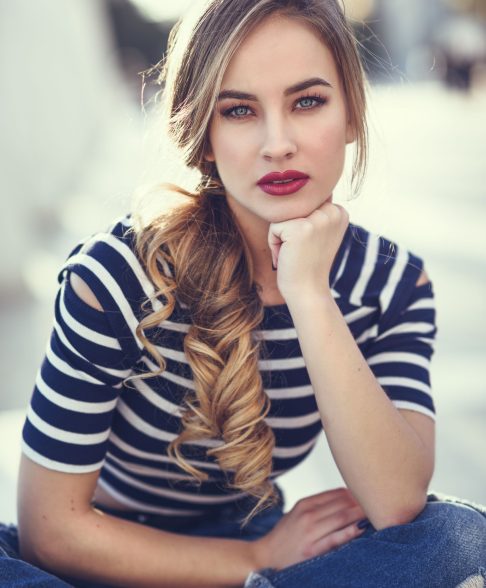 Blonde woman, model of fashion, sitting on a bench in urban background. Beautiful young girl wearing striped t-shirt and blue jeans in the street. Pretty russian female with pigtail.